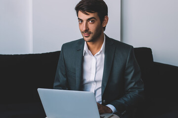 Positive young male entrepreneur sitting at cozy sofa and looking at camera in break of working on laptop device, prosperous man waiting for business meeting and preparing for presentation via netbook