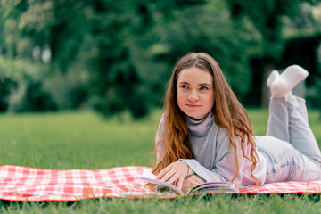 close up in the park on a red blanket girl lies on her stomach propping her head up reading a book...