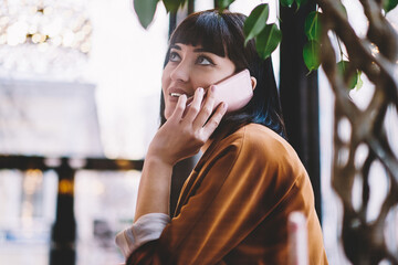 Positive smiling hipster girl calling to consultancy center using 4g connection on cellular phone while sitting at cafeteria with loft interior, concept of modern technology and communication