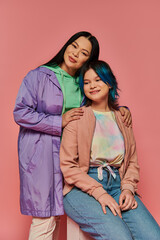 an Asian mother and her teenage daughter, sit side by side in casual attire against a vibrant pink background.