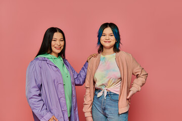 An Asian mother and her teenage daughter stand together in casual wear in front of a vibrant pink wall.