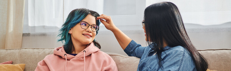 A mother with unique blue hair is tenderly combing her teenage daughters hair at home in a...