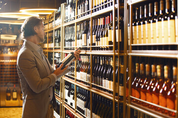 Professional sommelier holding bottle of wine in cellar, reading information on sticker about...