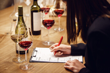 woman sommelier tasting red wine writing notes sitting at teh table being in cellar.