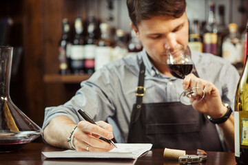 Male sommelier tasting red wine and making notes at bar counter