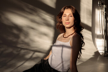 Naklejka premium Dramatic portrait of a young woman with long wavy hair, dressed in a white t-shirt and jeans, posing in the shadows and the sunset light.