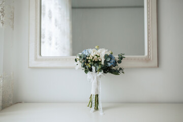 A bouquet of flowers sits on a table in front of a mirror. The flowers are blue and white, and the arrangement is simple and elegant. The mirror reflects the flowers, creating a sense of depth