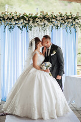 Fototapeta premium A bride and groom kiss in front of a blue curtain. The bride is wearing a white dress and the groom is wearing a suit