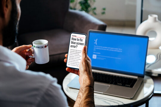 Man with mobile phone searching information to fix blue screen of death or BSOD of his laptop. Faulty operating system error.