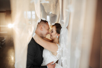 A bride and groom are hugging each other in front of a curtain. The bride is wearing a white veil and the groom is wearing a suit. Scene is romantic and intimate