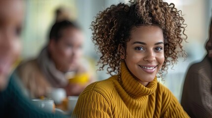 Obraz premium A joyful woman with curly hair smiles warmly while seated in a cozy cafe setting with friends in the background, creating a cheerful ambiance.