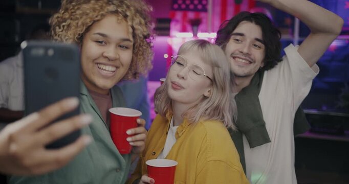 Carefree friends taking selfie posing for smartphone camera having fun with drinks chilling at night party. Self-portrait and modern technology concept.