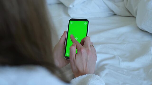 The blonde is sitting on the bed. Women's hands will scroll the phone with a green screen. 4k video. High quality 4k footage