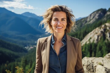 Naklejka premium Portrait of a cheerful woman in her 40s dressed in a stylish blazer on backdrop of mountain peaks