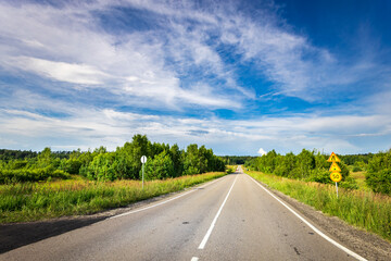 A paved country road winds its way through lush greenery, disappearing into the distance under a vibrant blue sky adorned with fluffy white clouds. A yellow speed limit sign marks the way.