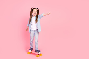 Full size photo of small girl dressed denim shirt pants riding skateboard to empty space raising fist isolated on pink color background