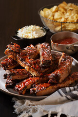 A plate of stacked ribs with side of BBQ sauce, coleslaw and potato chips