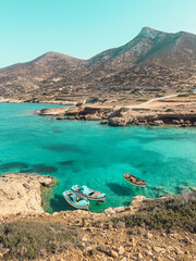 Three small boats gently float in the turquoise waters of a secluded cove in Donousa, Greece. Lush greenery contrasts with the brown cliffs in the distance