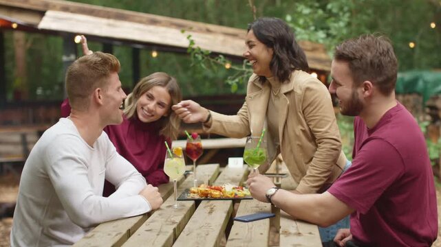 group of friends enjoys an outdoor picnic, sharing food and drinks. One woman playfully feeds a man while others smile and laugh