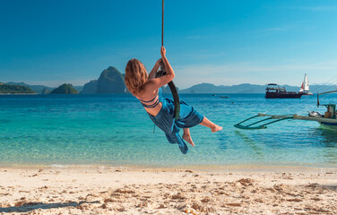 A woman with long blonde hair, wearing a blue dress, swings on a tire swing over crystal clear turquoise waters.