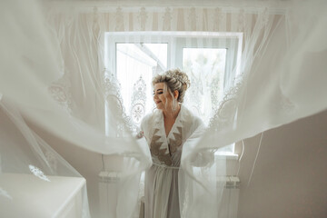 A woman in a white gown is standing in front of a window, with a white curtain draped over her. She...