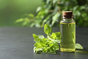 Essential oil in bottle and oregano twigs on dark textured table against blurred green background, closeup. Space for text