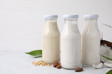 Different types of vegan milk in bottles and ingredients on white tiled table, space for text