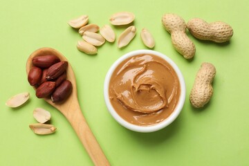 Tasty peanut butter in bowl and spoon with groundnuts on green table, flat lay