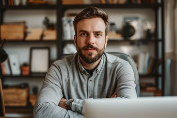 Obraz premium A man with a beard and a gray shirt is sitting in front of a laptop. He is looking at the camera with a serious expression