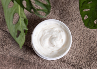  Opened cream jar on brown towel near green plant top view, everyday facial care