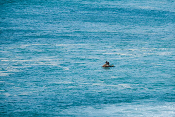 A lone surfer sitting on a rock in the ocean, surrounded by blue waters and the distant horizon,...