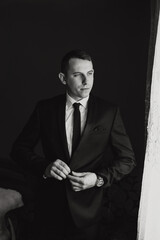 A man in a suit is standing in front of a window, adjusting his tie. Concept of formality and professionalism, as the man is dressed in a suit and tie