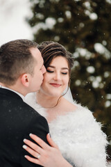 A bride and groom are embracing in the snow. The bride is wearing a white fur stole and the groom is wearing a black suit