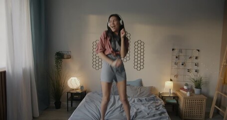 Joyful Asian woman listening to music with headphones and dancing on bed having fun in bedroom in evening. Modern lifestyle and entertainment concept. - Powered by Adobe