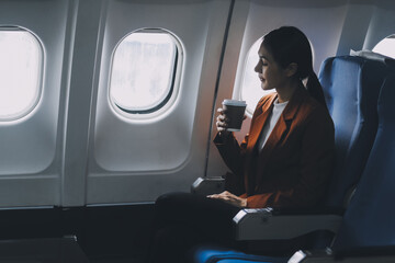 Fototapeta premium Young Asian female tourist traveling alone on a plane is drinking hot coffee and looking out the window.