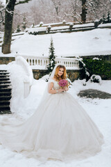 A woman in a white wedding dress is standing in the snow holding a bouquet. The scene is set in a garden with a white fence and a white staircase