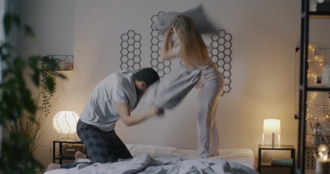 Man and woman happy young couple fighting pillows and laughing having fun in bed in evening. Romantic relationship and family lifestyle concept.