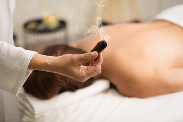 Female hand with smoking Palo Santo stick. Massage therapist using aroma at work with client lying face down in salon, close-up, selective focus.