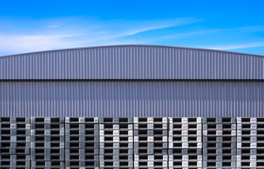 Row of many plastic pallets stacked in front of gray aluminium warehouse building wall against blue sky background