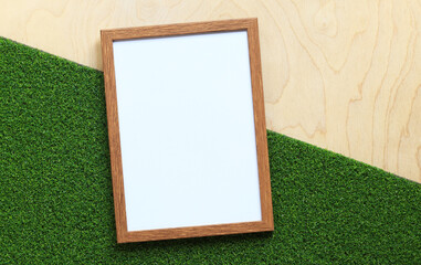 Blank frame with white sheet on light wooden background with green artificial grass. Simple wooden frame. Place for text. Blank photo with copy space. Blank frame for photo or text