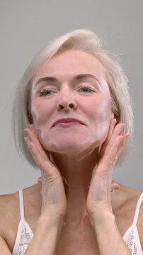 Close up Portrait: old woman touching facial skin and head, smiles. Senior female in the white studio. Beauty concept, skincare treatment, cosmetics and anti age plastic surgery.
