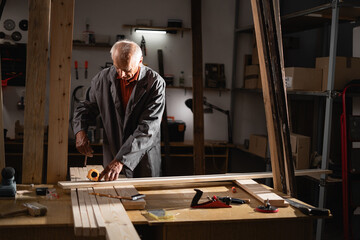 Old carpenter measuring the length of a board for future cutting, using a tape measure, working in his workshop in the evening