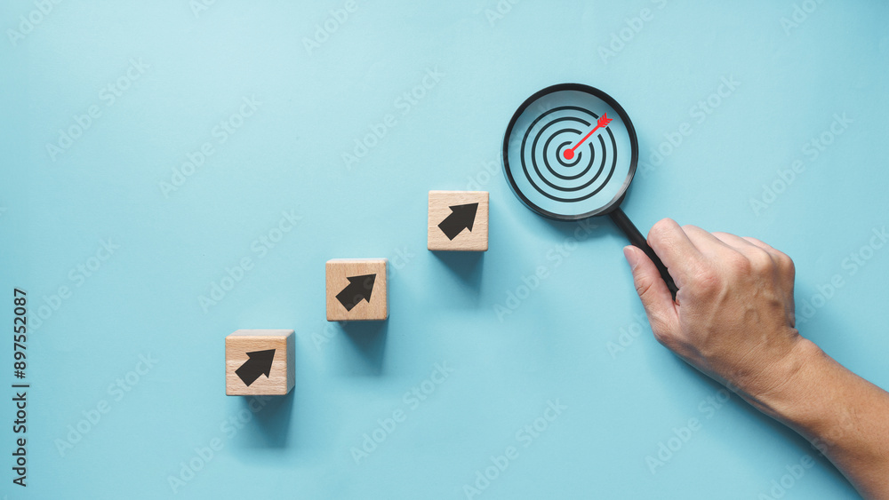 Poster a hand holding magnifying glass over wooden blocks with arrows point upward to red dartboard with ar