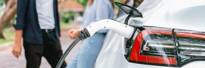 Panorama banner of lovely young couple recharging battery for electric car during autumnal road...