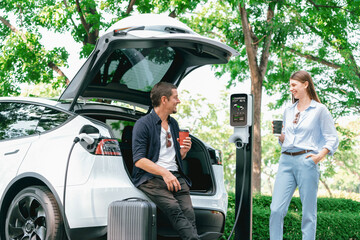 Fototapeta premium Lovely young couple drinking coffee while recharging battery for electric car during road trip travel EV car in natural forest or national park. Eco friendly travel during vacation and holiday. Exalt