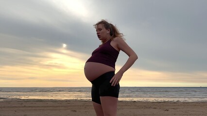 Young pregnant active woman doing exersice outdoors, stratching her body and meditate on the sea, beach at sunset in the evening