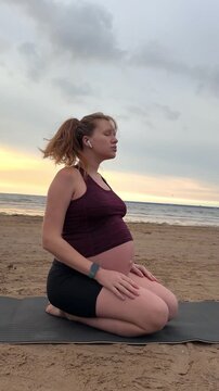 Young pregnant active woman doing exersice outdoors, stratching her body and meditate on the sea, beach at sunset in the evening
