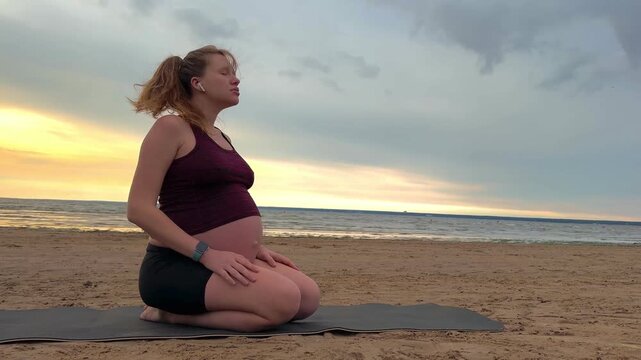 Young pregnant active woman doing exersice outdoors, stratching her body and meditate on the sea, beach at sunset in the evening
