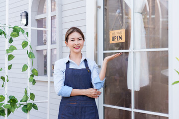 Beautiful Asian girl small business spa owner presenting open sign at door. Wearing blue apron and light blue shirt, smiling, welcoming customers. Professional, inviting, clean spa environment.