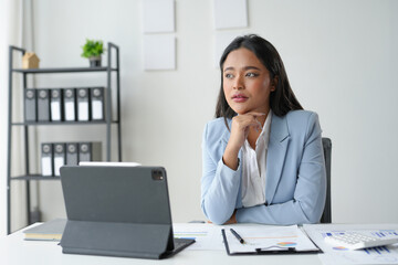 Young asian businesswoman is looking away and thinking about a new project while sitting at her desk in a modern office